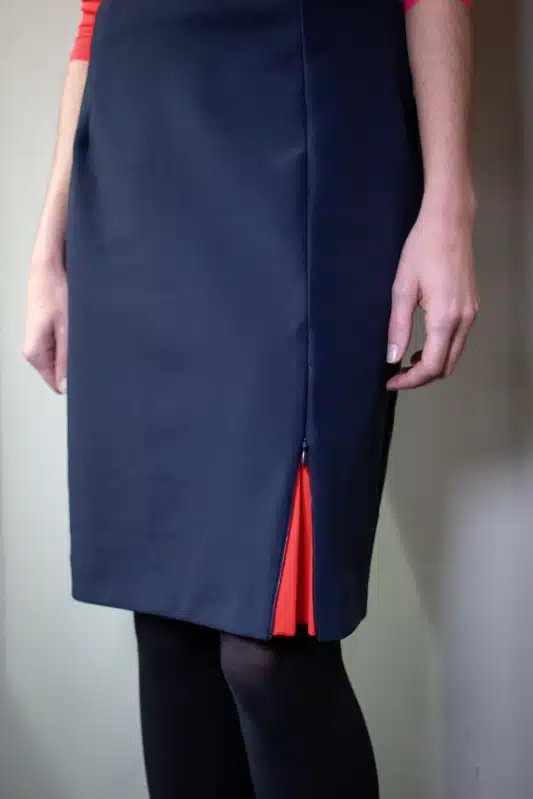 vue partielle d'une femme debout, portant une jupe marine s'ouvrant au bas de la jambe gauche sur une quille corail