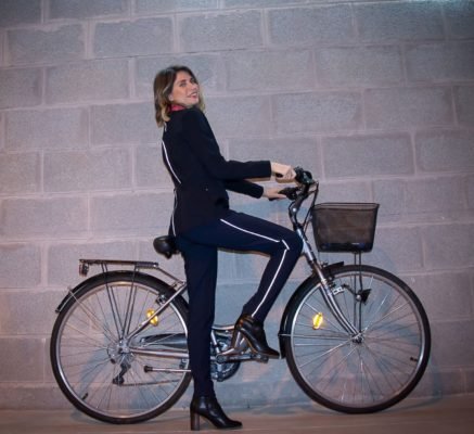 INSTA_SMOKING_VELO_3_gauche_à _droite pour bouger chic, jeune femme en pantalon marine et veste noire Cyclamelle, dont les passepoils éclairés renvoient la lumière, elle tient le guidon de son vélo, un pied posé sur la pédale
